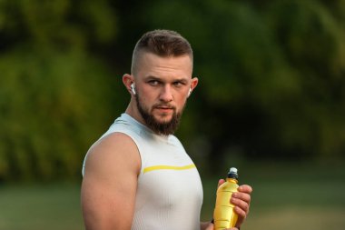 Genç ve zinde bir adam kablosuz kulaklıkla müzik dinliyor ve içme suyu içiyor ya da yaz parkında spor yaptıktan sonra biberondan beslenme suyu içiyor, aktif ve sağlıklı bir yaşam tarzı. Yüksek kalite fotoğraf