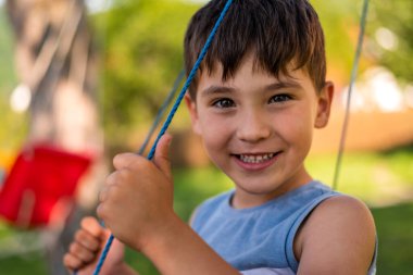 Mutlu bir çocuk yaz parkında salıncakta sallanırken kameraya bakıp gülümsüyor. Yüksek kalite fotoğraf