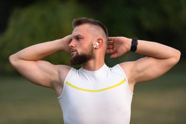 Koşmadan ya da parkta egzersiz yapmadan önce esneme egzersizleri yapan genç bir adam motive olmak için kablosuz kulaklıkla müzik dinler. Aktif yaşam tarzı, fitness ve açık hava egzersiz konsepti