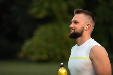 Genç, zinde bir adam kablosuz kulaklıklardan müzik dinler ve bir yaz parkında spor yaptıktan sonra dinlenir. Aktif ve sağlıklı bir yaşam tarzı sergiler. Yüksek kalite fotoğraf