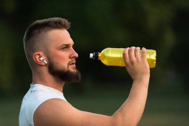 Genç ve zinde bir adam kablosuz kulaklıkla müzik dinliyor ve içme suyu içiyor ya da koşudan sonra bir şişeden beslenme suyu içiyor ya da parkta spor yapıyor, aktif ve sağlıklı bir yaşam tarzı. Yüksek kalite fotoğraf