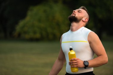 Gözleri kapalı, kablosuz kulaklıkla müzik dinleyen ve parkta koşudan ya da antrenmandan sonra dinlenen sağlıklı yaşam tarzı, fitness ve açık hava egzersizi konsepti olan genç bir adam. Yüksek kalite