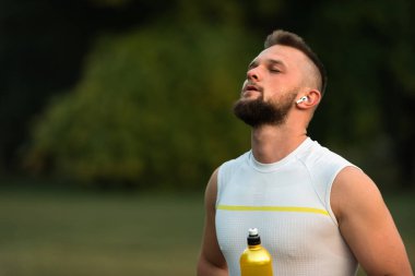 Gözleri kapalı, kablosuz kulaklıkla müzik dinleyen ve parkta koşudan ya da antrenmandan sonra dinlenen sağlıklı yaşam tarzı, fitness ve açık hava egzersizi konsepti olan genç bir adam. Yüksek kalite