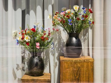 Güneş ışığını yansıtan ve pencerenin kenarındaki rahat köşeye biraz cazibe ve doğal zarafet katan canlı yaz çiçekleriyle dolu iki kırsal siyah vazo. Yüksek kalite fotoğraf