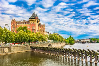 A picturesque view of Prague along the Vltava River, showcasing elegant traditional buildings lining the streets, bathed in warm, idyllic colors and set against a brilliant blue sky with soft clouds. clipart