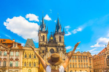 Kollarını kaldırmış turist kadın Prag 'ın tarihi şehir meydanında güneşli mavi gökyüzünün altındaki renkli binalarla çevrili Tyn' in önünde Meryem Ana 'nın gotik kilisesine bakıyor.