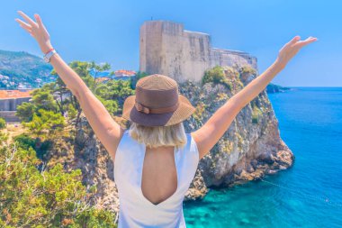 Kameraya arkası dönük duran turist kadın, kollarını kaldırmış, Fort Lovrijenac 'a tepeden bakan taş duvarlar ve Adriyatik Denizi' nin arka planında tarihi Dubrovnik kasabası