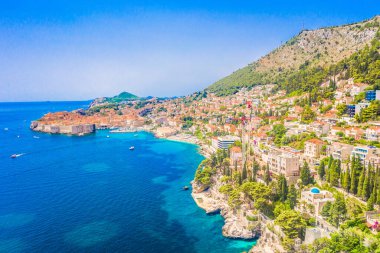 Dubrovnik kıyı şeridinin insansız hava aracı görüntüsü kayalık kıyıları, mavi Adriyatik suları ve parlak yaz gökyüzünün altında UNESCO tarafından listelenmiş tarihi eski kasaba