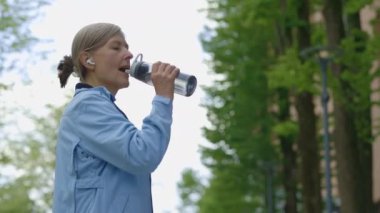 Eğitimden sonra spor şişesinden su içen olgun bir kadının arka planda susadığını hissedişinin yan görüntüsü. Spor ve sağlıklı yaşam tarzı kavramı