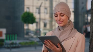 Ofis merkezine yakın oturan, dışarıda çalışan, iş için tablet kullanan tesettürlü mutlu Arap iş kadını. İş merkezi arka planında tablet üzerinde çalışan kadın yönetici