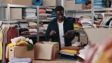 Afroamerikan erkek gönüllü depoda durup tablet kullanarak bağış için kıyafet almayı düşünüyor. Arka plandaki raflara aitti. Bağış ve insani yardım kavramı