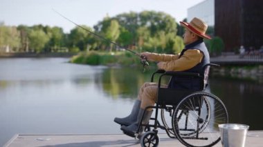 Tekerlekli sandalyede oturan engelli Asyalı adamın şapkası ve özel balıkçı kıyafetleri yerel göldeki iskeleden olta kamışının yan görüntüsü. Engelliler ve balıkçılık kavramı