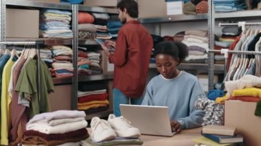 İkinci el bilgisayar kullanan çok ırklı bir kadın ya da arka planda birçok eşyası olan bir giyim merkezini yeniden kullanmak. Arka plandaki erkek arkadaşı. Sürdürülebilirlik kavramı