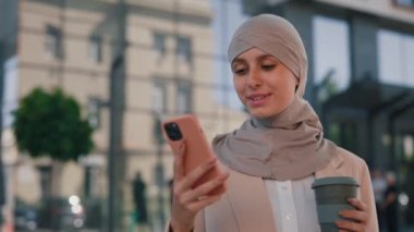 İş merkezinin yanında tesettürlü gülümseyen Arap iş kadını akıllı telefon kullanarak elinde kahve fincanıyla arkadaşlarıyla mesajlaşıyordu. İnsanlar ve teknoloji sektörü kavramı