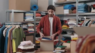 Erkek işçi eşyaları kutuya koyar ve ikinci el çalışır ya da arka planda birçok eşyayla birlikte kıyafet merkezini yeniden kullanır. İnsanlar, gönüllü, ikinci el, bağış konsepti