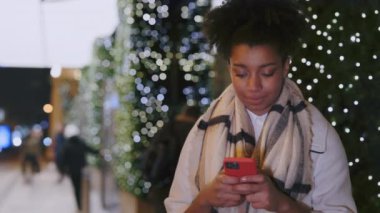 Noel arifesinde alışveriş merkezinin yakınında duran akıllı telefonu kullanan Afrikalı Amerikalı kadın. Gülümseyen kadın mesajları. Eğlence sezonu