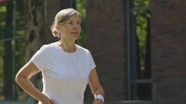Πλευρική Άποψη Του Χαμογέλασε Ώριμη Γυναίκα Τρέχει Και Φορώντας Ακουστικά — Αρχείο Βίντεο