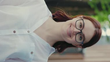 İş merkezinin yakınında kameraya gözlüklü, resmi takım elbiseli, gülümseyen kızıl iş kadınının dikey video resmi. Halk ve iş kavramı