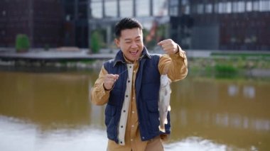 Asyalı erkek kendini mutlu hissediyor çünkü bir balık yakaladı ve oltaya takılı tutuyor ve kameraya bakıyor. İnsanlar ve balıkçılık konsepti