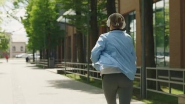 Arka planda, spor giysiler giymiş, kulaklık takmış, sokakta koşan yaşlı bir kadın var. Spor ve sağlıklı yaşam tarzı kavramı. Gençlik Yaşlanma.