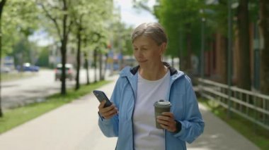Sokakta koşan ve elinde kahve fincanıyla akıllı telefon kullanan sportif bir koşucu kadın. Spor ve sağlıklı yaşam tarzı kavramı. Gençlik Yaşlanma