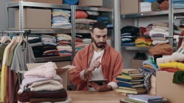 Erkek gönüllü, bağış için kıyafet düşünürken bir yandan da tablet kullanırken depoda oturuyor. Arka plandaki raflara aitti. İnsani yardım kavramı