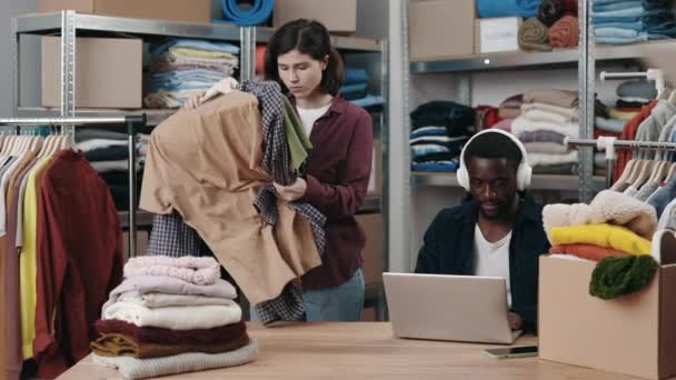 不同的人在送货仓库工作 分拣捐赠的衣服 白人女工看着衣架 而她的男同事则用手提电脑和耳机 — 图库视频影像