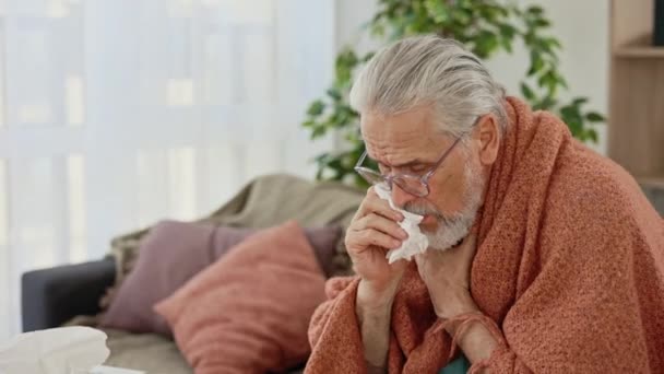 体弱多病的老人披着温暖的毯子 咳嗽不舒服的老爷爷坐在沙发上 感觉得了重感冒病毒的症状 老年男性有健康问题共19种流行观念 — 图库视频影像
