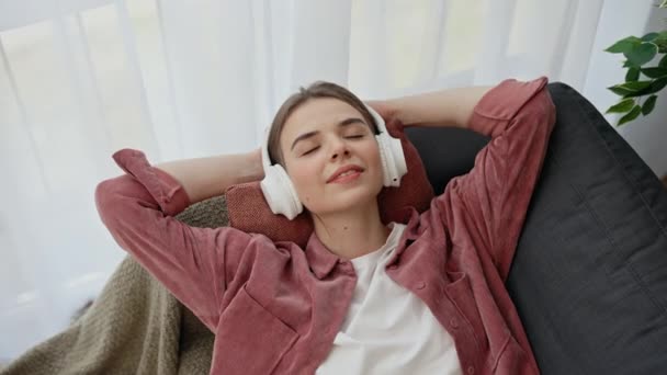 宽松的高加索女人躺在家里的沙发上 喜欢用耳机听音乐 人与技术概念 — 图库视频影像