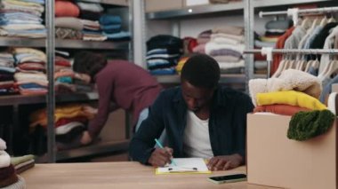 Çok ırklı bir adam, beyaz kadın meslektaşı elbiselerini masaya koyarken ve onları ayırmaya hazırlanırken not alıyor. Merkezi ve ikinci el kavramını yeniden kullan