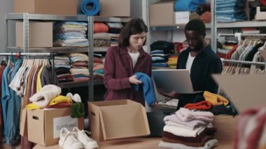 Çok ırklı engelli erkek gönüllü, bağış merkezinde bir sürü kıyafetle birlikte dizüstü bilgisayar kullanıyor. Meslektaşı kutuya koyuyor. İnsani yardım ve gönüllüler konsepti