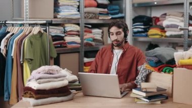 Çok kültürlü erkek gönüllü, kulaklık takıyor dizüstü bilgisayar kullanıyor ve yeniden kullanım merkezinde çalışırken müşteriyle sohbet ediyor. Bağış, hayır kurumu ve ikinci el konsepti