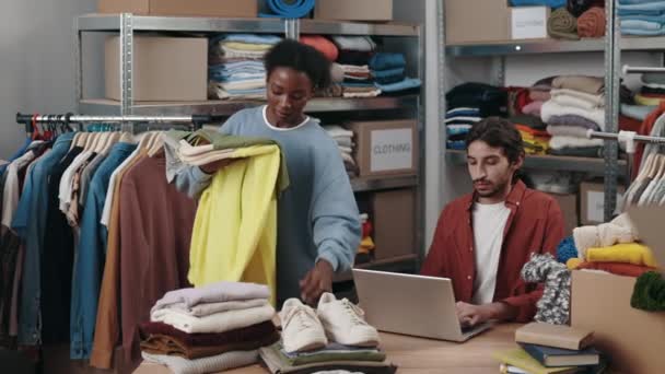 多种族女性在衣架上挑选衣服进行回收或捐赠 而男性同事则在慈善商店使用笔记本电脑 重复使用 有意识的消费者 可持续性概念 — 图库视频影像