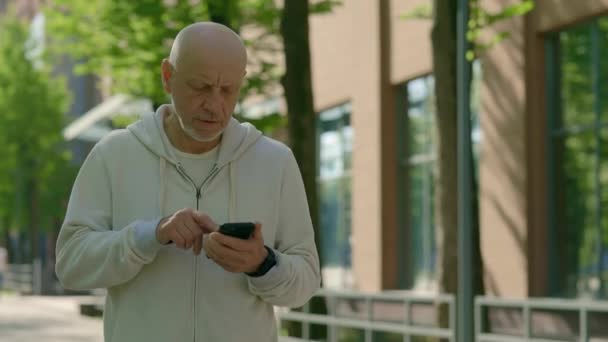 老人は通りの背景にスマートフォンを使って通りを通っています 成熟した男性アスリートは 電話メッセージを読むか アプリをダウンロードします スポーツ 健康的なライフスタイルのコンセプト — ストック動画