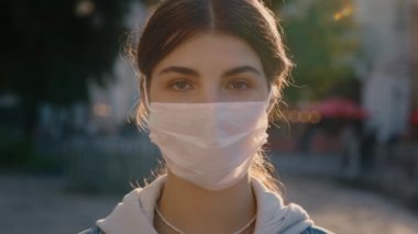 Koruyucu tıbbi maske takan ve sokakta dikilen güzel siyah saçlı genç bir kadının portresi. Güvenli ve Mutlu Kadın Sosyal Mesafe Uygulaması, Karantina. Bulanık Şehir Arkaplanı