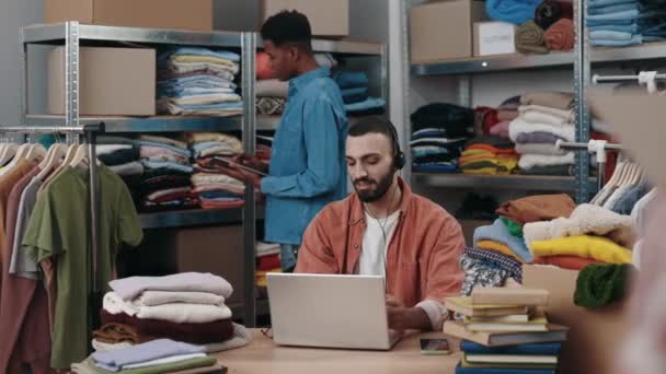 ハッピーディセーブル男性マネージャーは ヘッドセットでクライアントと話をし 衣料品店でラップトップで働いています 彼の同僚は背景にタブレットを使っている 小さな仕事だ 自営業員 — ストック動画