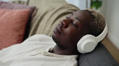 Genç ve yakışıklı Afro-Amerikalı adamın evdeki koltuğunda oturup, zor bir günün ardından beyaz kulaklıklarla müziğin keyfini çıkarmasını. Teknoloji ve iletişim konsepti.