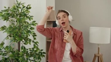 Genç beyaz kadın evde şarkı söylerken elinde telefonuyla müzik dinliyor ve dans ediyor. Kameraya şarkı söyleyen ve rahat bir dairede dans eden mutlu beyaz kadın.