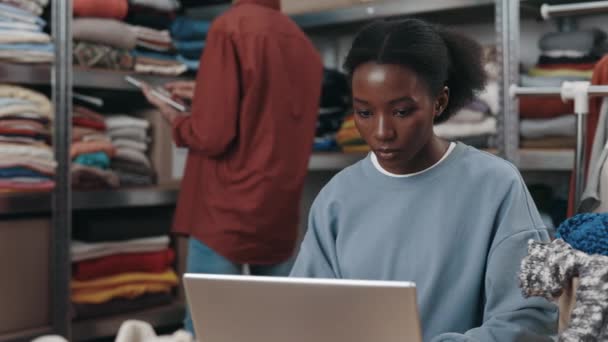 多种族妇女坐在笔记本电脑前 在仓库里工作 背景上有很多东西 她在幕后工作的男同事 — 图库视频影像