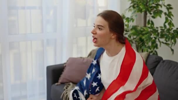 白人妇女在家里为她最喜欢的足球队加油加油加油 兴奋的年轻女球迷裹着美国国旗观看电视转播的国际比赛 体育和娱乐概念 — 图库视频影像