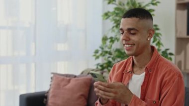 Kanepede oturan ve akıllı telefon kullanan mutlu çoklu etnik adama yakından bakın. Genç Afrikalı Amerikalı erkek öğrenci sosyal medyada sohbet ediyor. Teknoloji ve iletişim konsepti