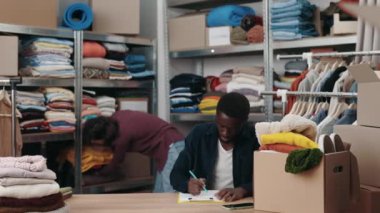 Çok ırklı bir adam, beyaz kadın meslektaşı elbiselerini masaya koyarken ve onları ayırmaya hazırlanırken not alıyor. Merkezi ve ikinci el kavramını tekrar kullan. Küçük işletme