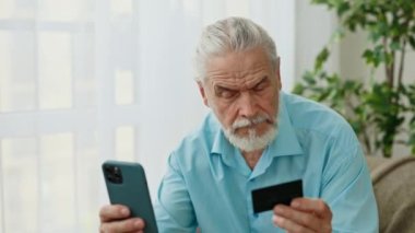 Evde oturan, günlük kıyafetlerini giymiş, online alışveriş için akıllı telefon ve kredi kartı kullanan, ödeme için veri giren, internet üzerinden alışveriş yapan ya da sipariş veren olgun beyaz bir adam.