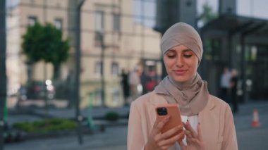 Güzel, gülümseyen Arap iş kadını resmi kıyafetler içinde tesettüre girmiş akıllı telefon kullanarak iş merkezinin yanında duruyor. İnsanlar ve teknoloji sektörü kavramı