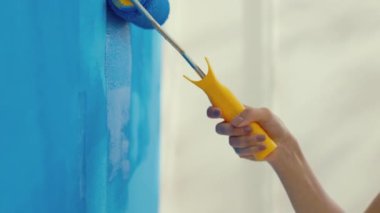 Kadın elleri boya silindirini tutuyor ve yeni dairenin duvarını mavimsi renklerle boyuyor. Paten duvara boya sürer. Aletleri kapatın. İnşaat, onarım ve yenileme