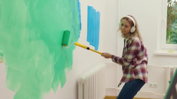 白人女性労働者は壁を塗装し イヤホンを使用して改装中に自宅で音楽を楽しんで踊ります 仕事中に音楽を聴いている面白い女性 国内活動概念 — ストック動画