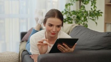 Genç beyaz kadın kanepede uzanmış, sohbet etmek için tablet kullanıyor ya da sosyal medya için. Evde alet kullanan mutlu genç bir kadın. Teknoloji ve iletişim konsepti