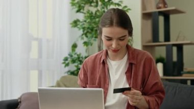 Kafkasyalı genç kadın evde oturuyor, günlük kıyafetlerini giyiyor, online alışveriş için laptop ve kredi kartı kullanıyor, ödeme için veri giriyor, online işlem veya sipariş veriyor.