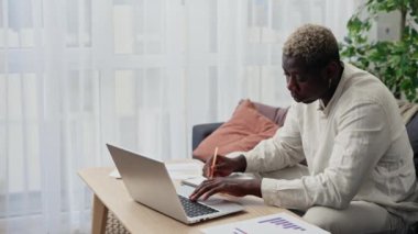 Genç Afro-Amerikalı adam evde çalışıyor, salgın sırasında internetten çalışıyor. Afrikalı Amerikalı adam internette dizüstü bilgisayar kullanarak ders çalışıyor ve notlar alıyor. Çevrimiçi eğitim kavramı