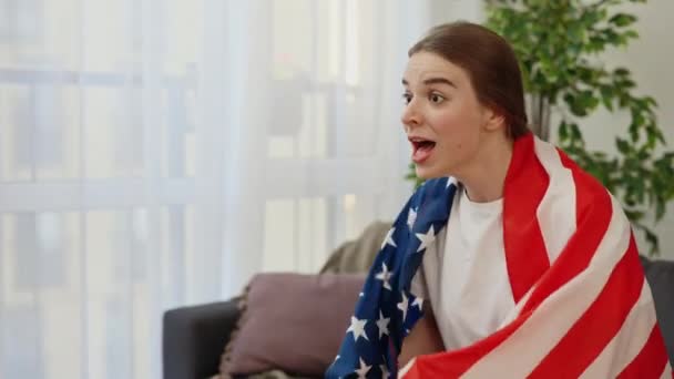 年轻的高加索女人在家里为她最喜欢的运动队加油加油 兴奋的年轻女球迷裹着美国国旗观看电视转播的国际比赛 体育和娱乐概念 — 图库视频影像